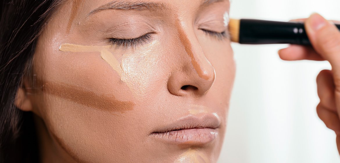 Junge Frau trägt mit ihrem Zeigefinger vor einem Spiegel Concealer unter ihren Augen auf.